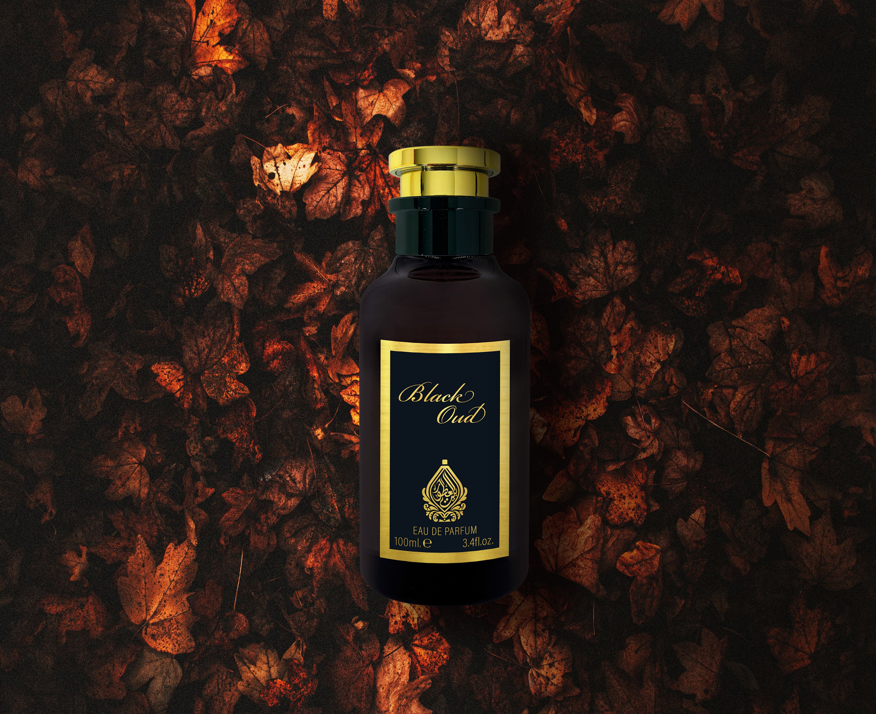 A bottle of Black Oud perfume lying on a surface with pine needles scattered around, with a focus on the perfume bottle that has a dark amber liquid inside.